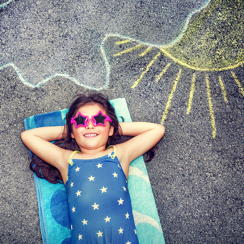 Happy little girl wearing swimsuit and stylish sunglasses lying down on the asphalt near picture of the sun comes out from behind the clouds, cute baby needs of summer holidays