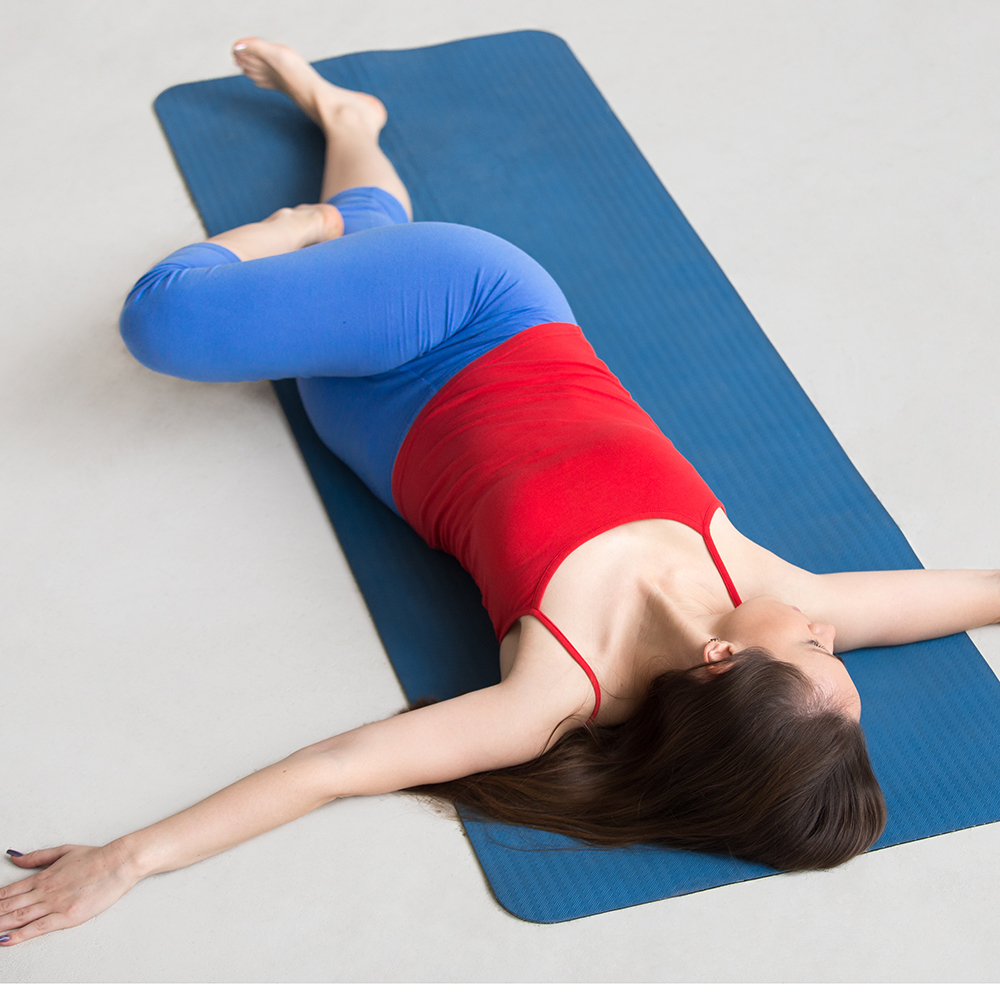 Beautiful happy young woman in bright colorful sportswear working out indoors on blue mat. Girl lying in Reclining Spinal Twist Jathara Parivartanasana resting after practice full length top view