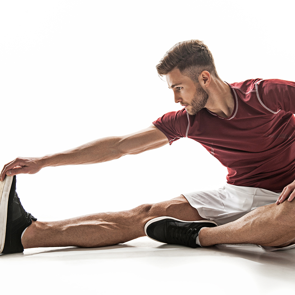 Fit young man is doing exercise with concentration. He is stretching hand to foot while sitting on floor. Isolated