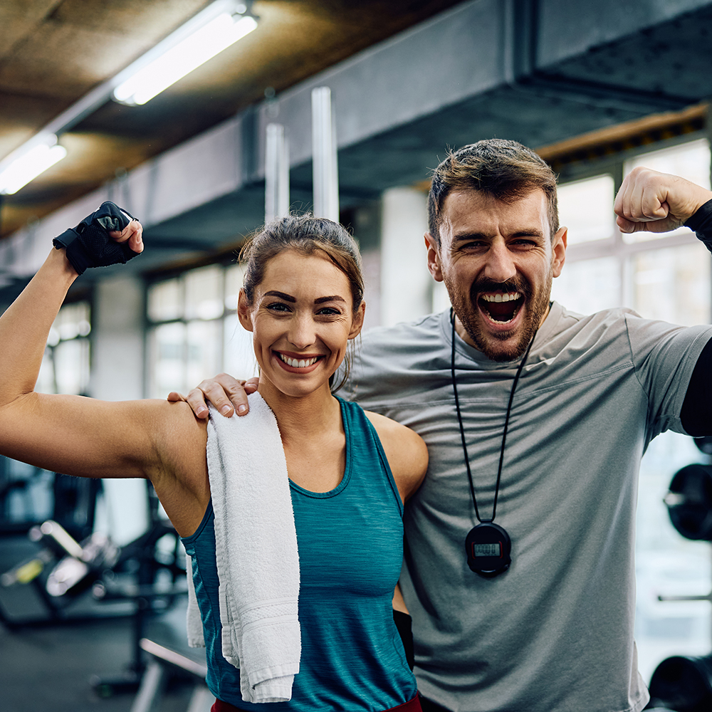 Happy athletic woman and her fitness instructor flexing muscles in a gym while looking at camera.