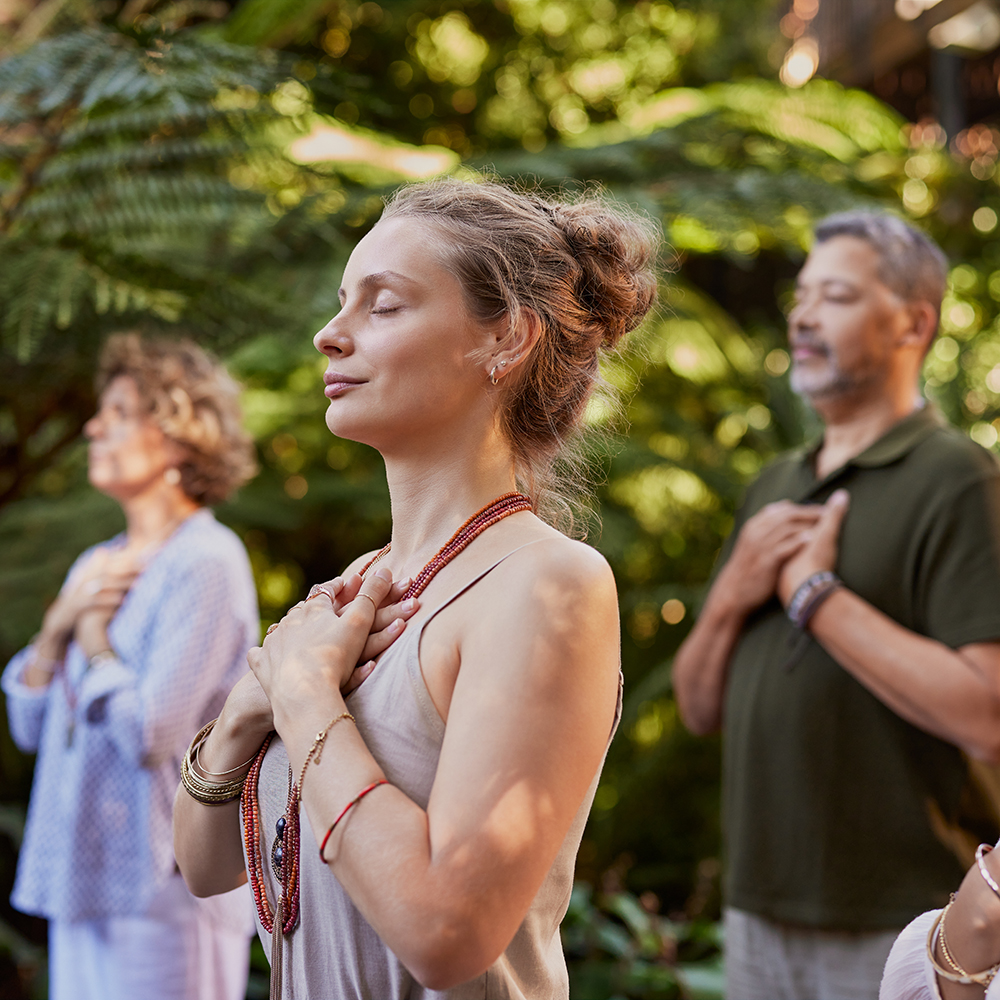 Multiethnic group of people standing outdoors with hands on their hearts, practicing mindfulness in a wellness retreat. Young woman in a wellness retreat center connecting to inner peace during a healing session. People doing spiritual practice feeling peace of mind and gratitude.