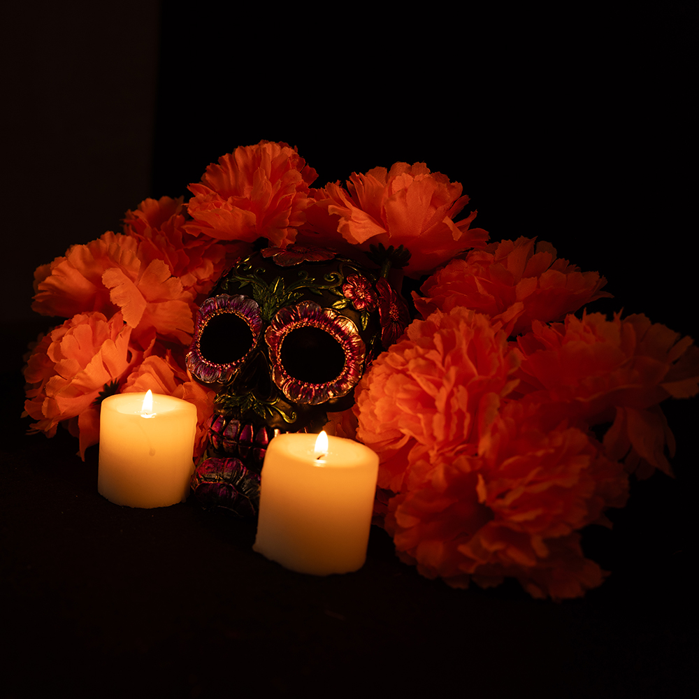A black skull adorned with metallic flowers, surrounded by a vibrant orange marigold wreath. Flanked by lit candles on a black background, capturing the essence of Mexico's Day of the Dead celebration